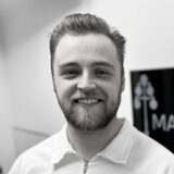 Person smiling warmly in a black and white photo, wearing a light-colored shirt, with a blurred background featuring an indistinct sign.