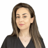 A woman with long, straight hair and a subtle smile wears a black top. She stands against a plain black background.