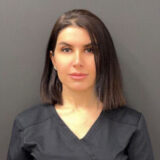 A young woman wearing a black medical scrubs top poses in front of a dark background, looking confidently at the camera.