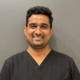 A smiling individual wearing dark scrubs stands against a neutral background, conveying a friendly and professional demeanor, likely representing a healthcare or dental setting.