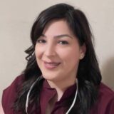 A smiling woman with shoulder-length dark hair is wearing a maroon top and has a stethoscope around her neck, appearing friendly and approachable.