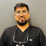 A smiling dental professional wearing black scrubs and glasses, with dental tools around his neck, stands against a plain gray background, conveying a friendly and approachable demeanor.