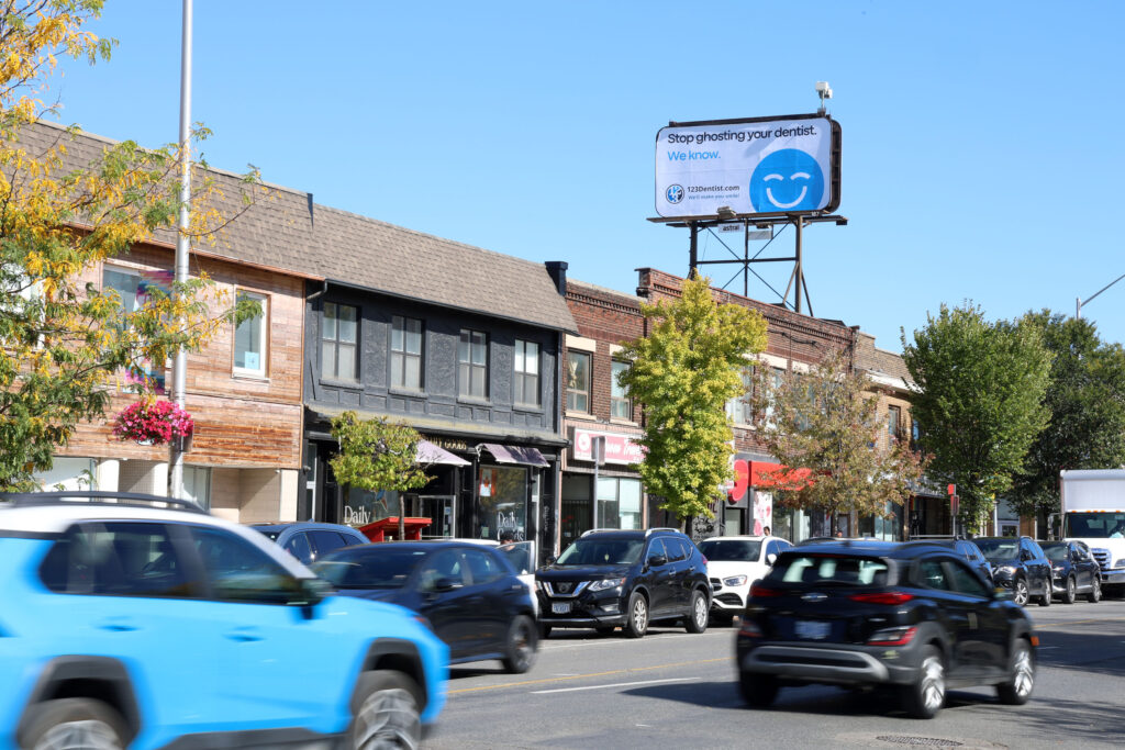 A bustling street scene features various shops and trees along the sidewalk. A billboard prominently displays a friendly blue smiley face, known as the 123Dentist Smiley, alongside a message promoting dental care. Cars fill the road, adding to the lively atmosphere.
