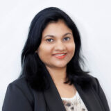 A woman with long black hair and a bright smile is wearing a black blazer over a light-colored top. She stands against a plain white background, exuding professionalism and warmth.