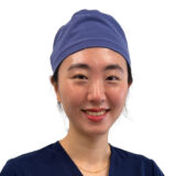 A young woman wearing a blue surgical cap and a navy scrubs top smiles warmly at the camera, conveying a friendly and professional demeanor.