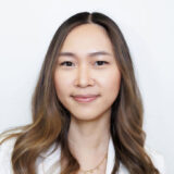 A smiling woman with long, wavy hair is portrayed against a plain white background. She is wearing a light-colored top and has a warm expression.