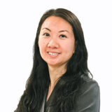 A smiling woman with long, dark hair poses confidently, wearing a gray blazer. She has a warm expression that conveys approachability and professionalism. The background is plain white, focusing on her friendly demeanor.