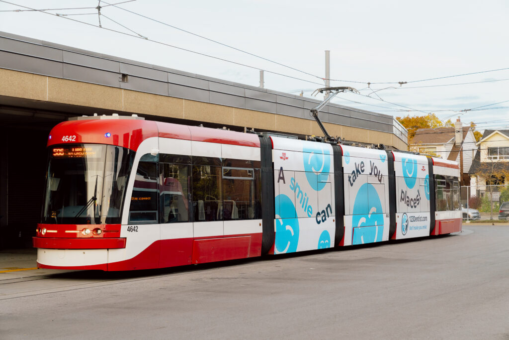 A modern tram with a red and white design is parked at a station. The side features advertisements, including a friendly blue smiley face, known as the 123Dentist Smiley, promoting dental hygiene. The setting captures an urban transport scene.
