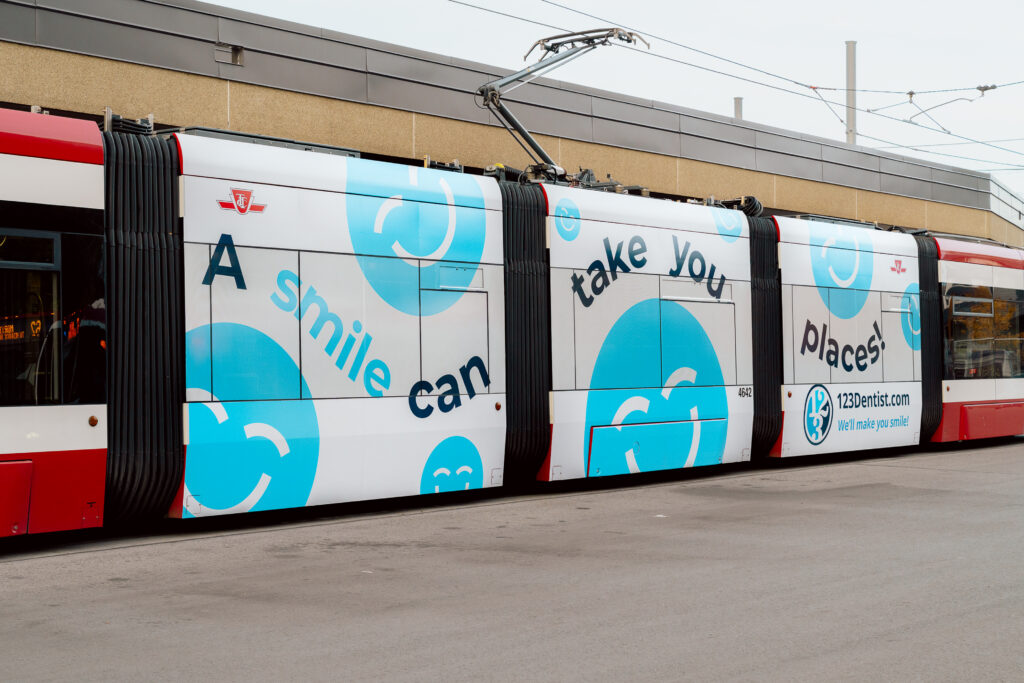 A tram features vibrant advertisements promoting dental health, displaying the slogan "A smile can take you places" alongside the cheerful 123Dentist Smiley. The design emphasizes the importance of smiles in a friendly and engaging manner.