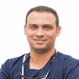 A smiling dental professional in scrubs stands confidently with arms crossed. He has short, dark hair and a friendly demeanor, embodying trust and approachability in a clinical setting.