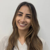 A smiling woman with long brown hair wears a light-colored medical scrubs top. She stands against a plain background, exuding warmth and professionalism.