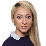 A woman with long, light blonde hair styled to one side smiles warmly at the camera. She wears a navy blue top with a collared shirt underneath, showcasing a professional yet approachable appearance.