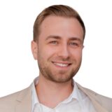 A smiling man in a light suit and white shirt is looking confidently at the camera. His short, styled hair complements his friendly expression, suggesting approachability and professionalism.