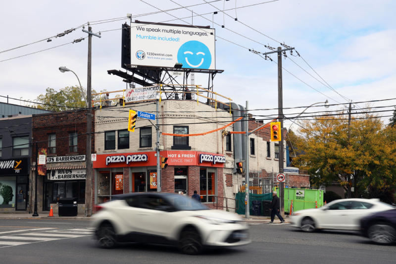 A corner street scene featuring a pizza shop with colorful signage. Above, a billboard displays the 123Dentist Smiley, promoting dental services. Cars are seen driving by on the busy road. Trees in the background hint at the autumn season.