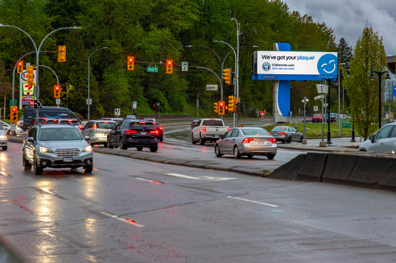 A busy intersection on a rainy day, featuring several cars stopped at traffic lights. A large billboard displays a friendly message from 123Dentist Smiley, promoting dental services. Lush green trees line the background, adding a touch of nature to the urban scene.