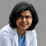 A smiling healthcare professional with shoulder-length dark hair and glasses wears a white lab coat over a blue shirt, posing against a neutral background.