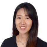 A woman with long dark hair smiling at the camera, wearing a black shirt and a necklace with a small heart pendant, set against a plain white background.