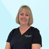 A smiling woman in black dental scrubs stands against a light blue background.