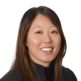 A woman with long brown hair is smiling against a white background, wearing a dark top.