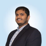 A smiling individual in a black suit and plaid shirt poses confidently against a light blue background.