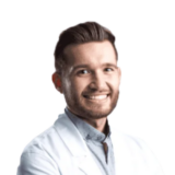 A smiling person with short hair and a beard, wearing a white coat, stands against a plain background.