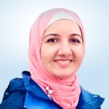 A smiling woman in a pink hijab poses against a light blue background, radiating warmth and positivity. She is wearing a blue jacket and appears approachable, embodying a friendly, welcoming demeanor.