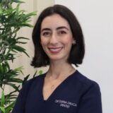 A smiling dentist poses in a professional setting, wearing a dark blue uniform with her name and title displayed. Green plants in the background add a touch of warmth to the environment.