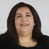 A woman with long dark hair and a black top smiles at the camera against a plain background.