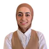 A person wearing a beige vest over a white shirt and a matching headscarf smiles at the camera against a plain white background.