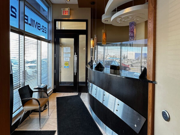 A modern dental office reception area with chairs, a curved front desk, decorative lights, and large windows.