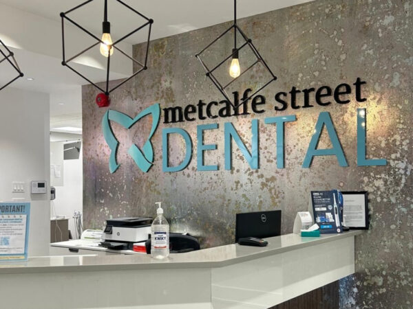 Reception area of Metcalfe Street Dental with a modern design, featuring a counter with informational pamphlets and a hand sanitizer bottle. The wall displays the dental office name in large letters, accented with hanging geometric light fixtures.