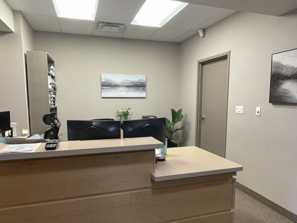 A well-lit dentist office reception area with a wooden desk, a computer, and a small plant. The walls display two black-and-white landscape photos, and a door is visible in the background.
