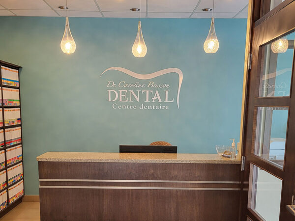 Dental office reception area with a sleek desk, light blue wall, and soft lighting from three hanging fixtures. The wall displays the dental clinic's logo. Shelves filled with files are on the left side. A glass door is partially open on the right.