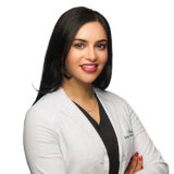 A confident woman with long dark hair wears a white lab coat over a black top and smiles with arms crossed.