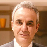 A man with short gray hair and a slight smile, wearing a suit, stands indoors with warm lighting in the background.