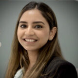 A person with long brown hair smiling, wearing a dark blazer and standing against a plain gray background.