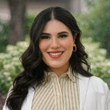 A smiling woman with long dark hair, wearing a striped shirt and a white coat, stands outdoors in front of greenery.