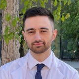 A person with short hair and a beard, wearing a white coat and dark tie, stands outdoors in front of a tree with green leaves.