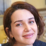 A woman with short brown hair smiles warmly at the camera. She is in a softly lit indoor setting.