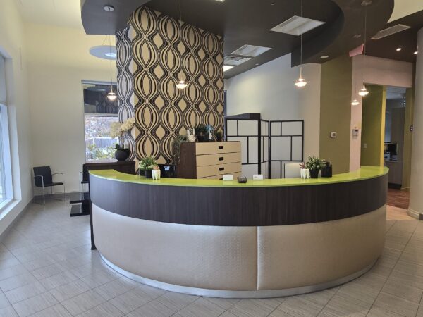 Modern dental office reception area with a curved desk, green accents, and decorative wallpaper. Large windows allow natural light to fill the space.