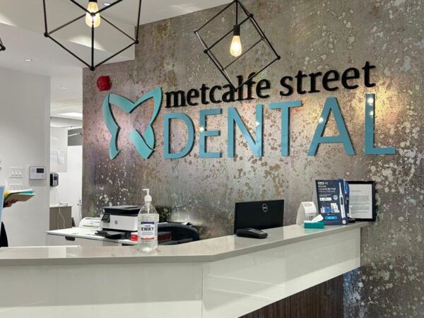 Dental office reception with a modern design, featuring a sign reading "Metcalfe Street Dental" against a textured wall. The desk displays hand sanitizer, a laptop, and office materials. A staff member is visible in the background holding paperwork. Hanging light fixtures add a touch of style.