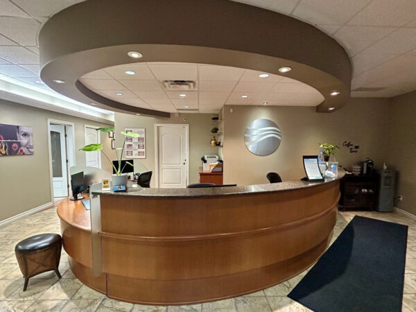 A modern dental office reception area with a wooden circular desk, overhead lighting, and seating. The decor features neutral tones and minimalistic design, creating a welcoming atmosphere.
