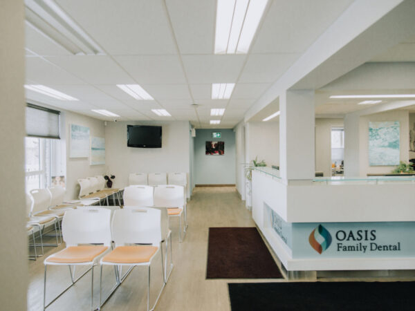A bright and clean dental office waiting area with rows of white chairs, a wall-mounted TV, and a reception desk labeled "Oasis Family Dental."