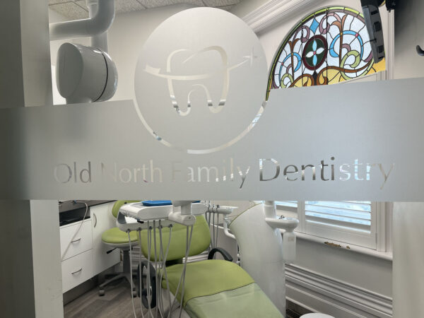 A dental office with frosted glass doors displaying a tooth logo and the name "Old North Family Dentistry." Inside, there are chairs and dental equipment, with a colorful stained glass window visible.