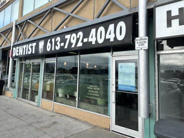Street view of a dentist's office with large windows and a sign displaying "DENTIST" and a phone number. The entrance door is on the right, and a security sign is on a post nearby.