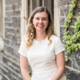 A woman with wavy blonde hair, wearing a white dress, smiling while sitting in front of an ivy-covered brick wall.