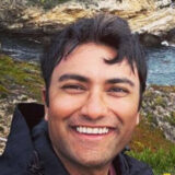 A person smiling outdoors with a rocky coastline and ocean in the background.