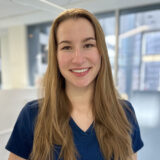 A smiling woman with long hair wearing a blue shirt stands in a bright office environment with modern glass windows in the background.