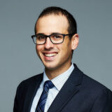 A person with short hair and glasses, wearing a suit and tie, smiles against a plain background.