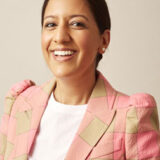 A person smiling warmly, wearing a pink and beige checkered blazer and a white top, with hair neatly pulled back.